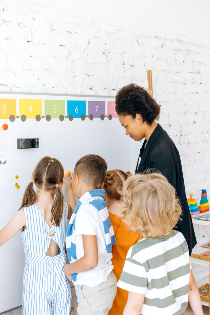 gallery-3 Children engaged in learning with a teacher at a colorful classroom whiteboard.
